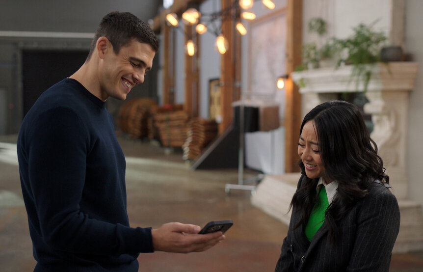 a smiling man holds his phone and another woman smiles at him