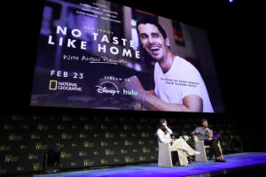 two people sit on stage with the no taste like home promo behind them