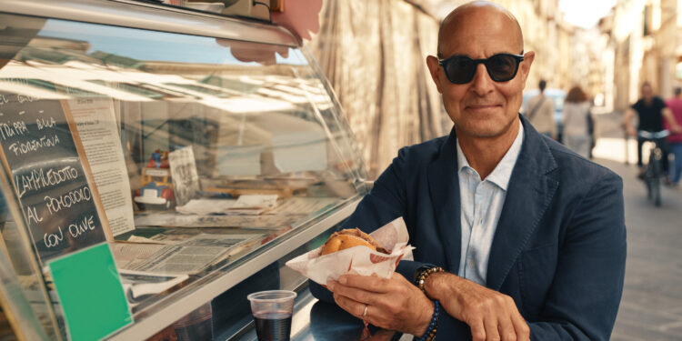 stanley tucci stands by a food stand in italy